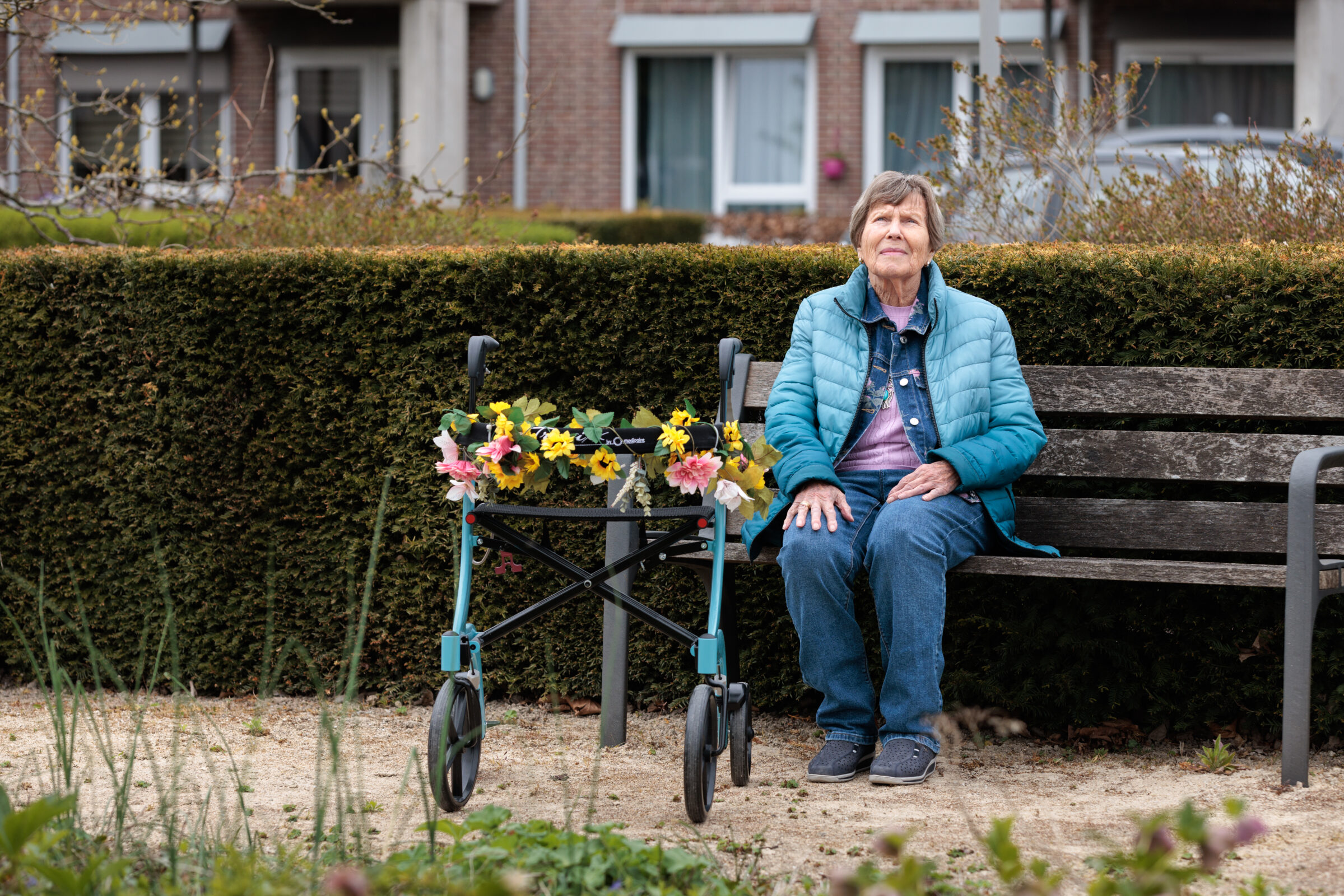 Een oudere vrouw zit vredig op een bankje in een tuin, naast een rollator versierd met levendige kunstbloemen.