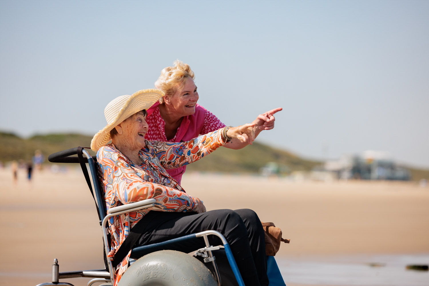 bewoner en medewerker zijn op het strand. Bewoner zit in een speciale strandrolstoel. Bewoner wijst richting de zee. Ze staan aan het begin van de zee.